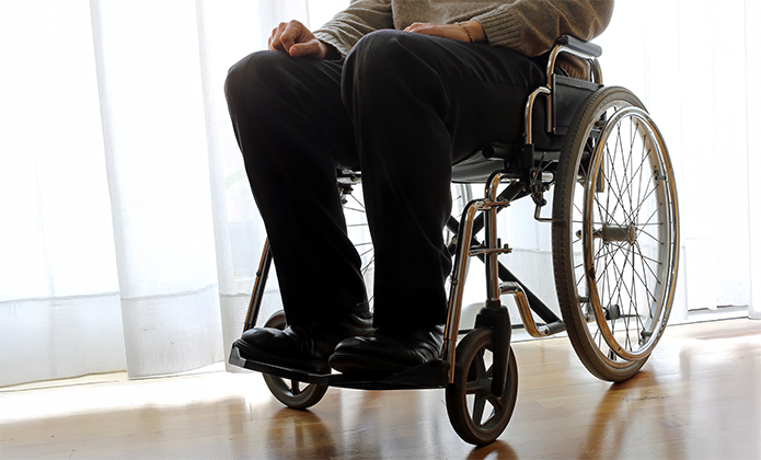 #ParaTodosVerem: A imagem mostra uma pessoa sentada em uma cadeira de rodas, fotografada da altura da cintura para baixo. A pessoa veste calças escuras e sapatos pretos, e um suéter cinza-claro na parte visível do tronco. As mãos repousam sobre o colo. A cadeira de rodas é manual, com estrutura metálica e aro grande visível à direita, refletindo luz. O ambiente tem piso de madeira clara e é iluminado por luz natural que entra através de uma cortina branca translúcida ao fundo, criando um clima suave e levemente difuso.